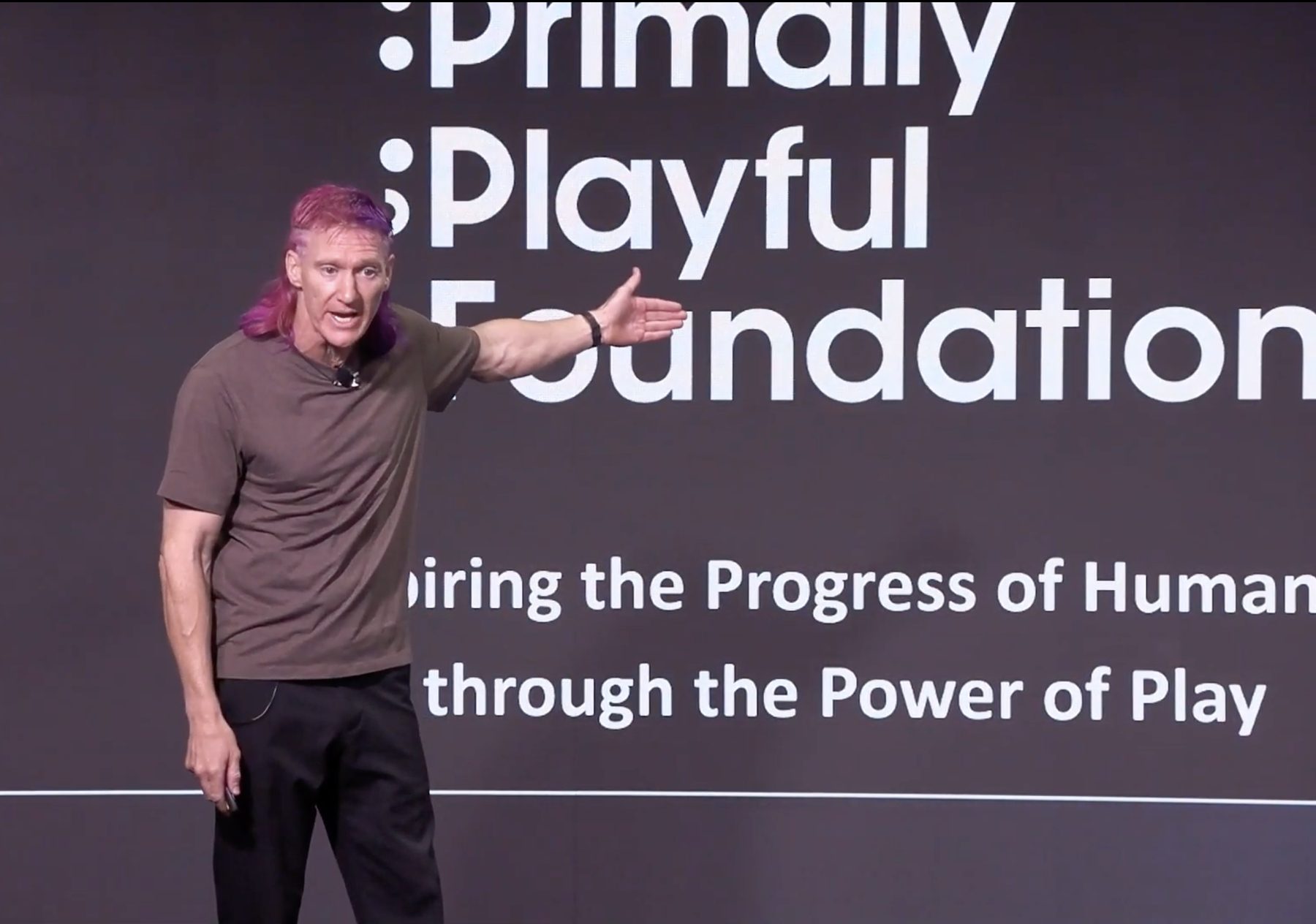 Jared Hippler on stage at Speak To Freedom, gesturing toward a screen showing the A Primally Playful Foundation logo